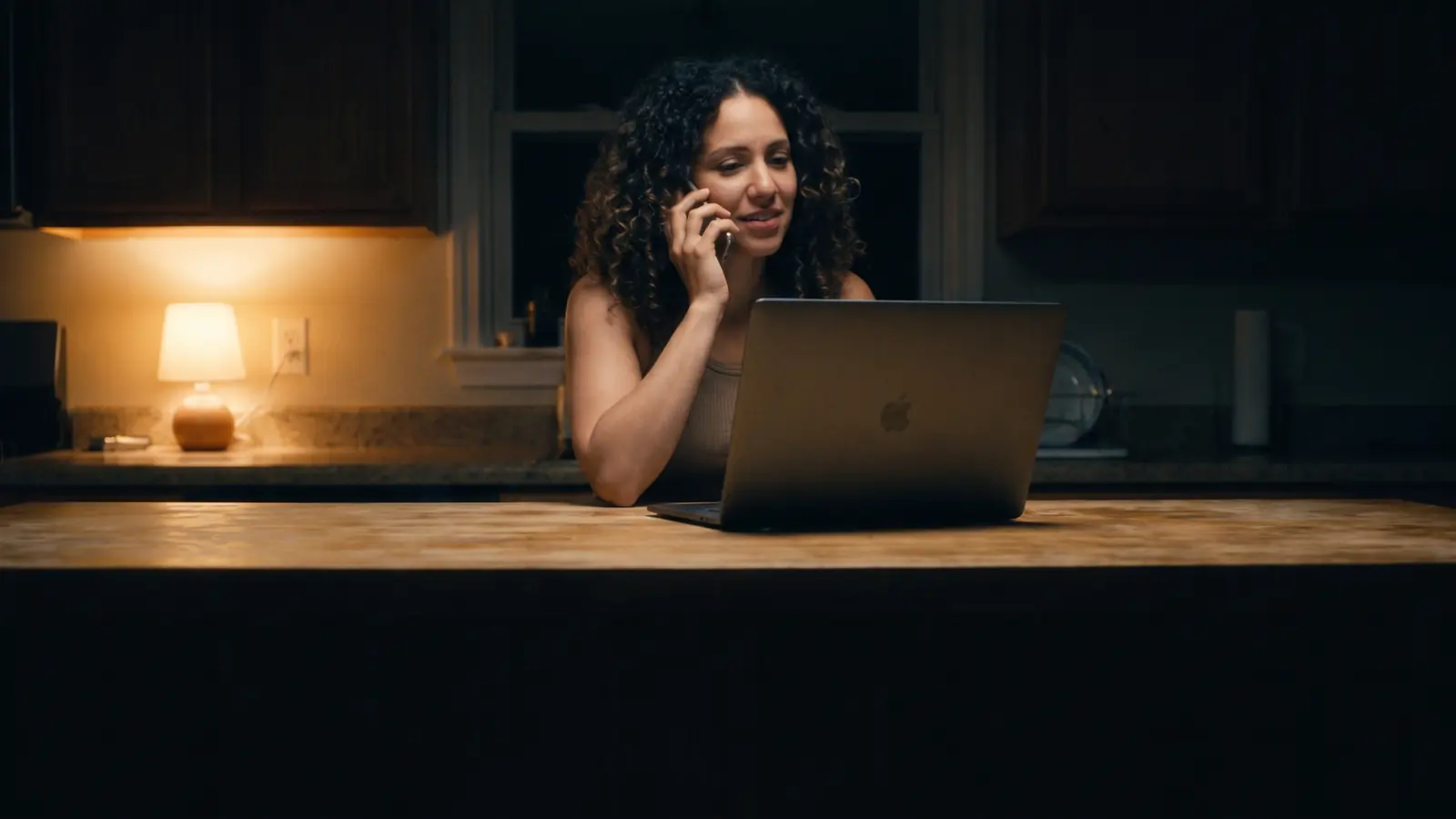 A woman on the phone at a kitchen island late in the evening, a laptop open in front of her.