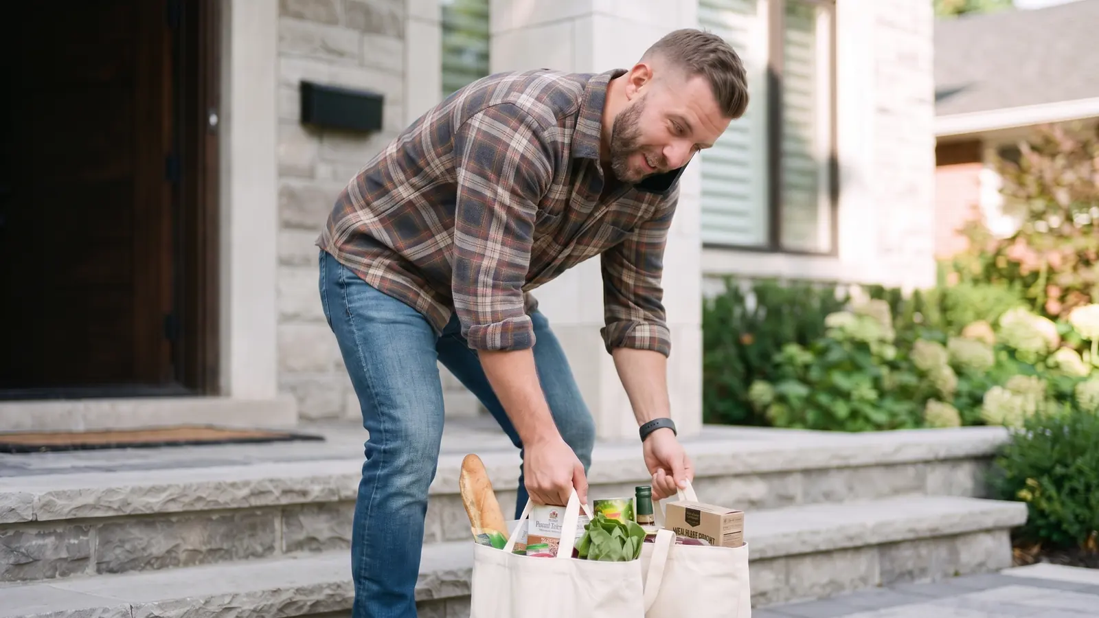 A man carrying groceries up his front steps, pausing to check his phone.