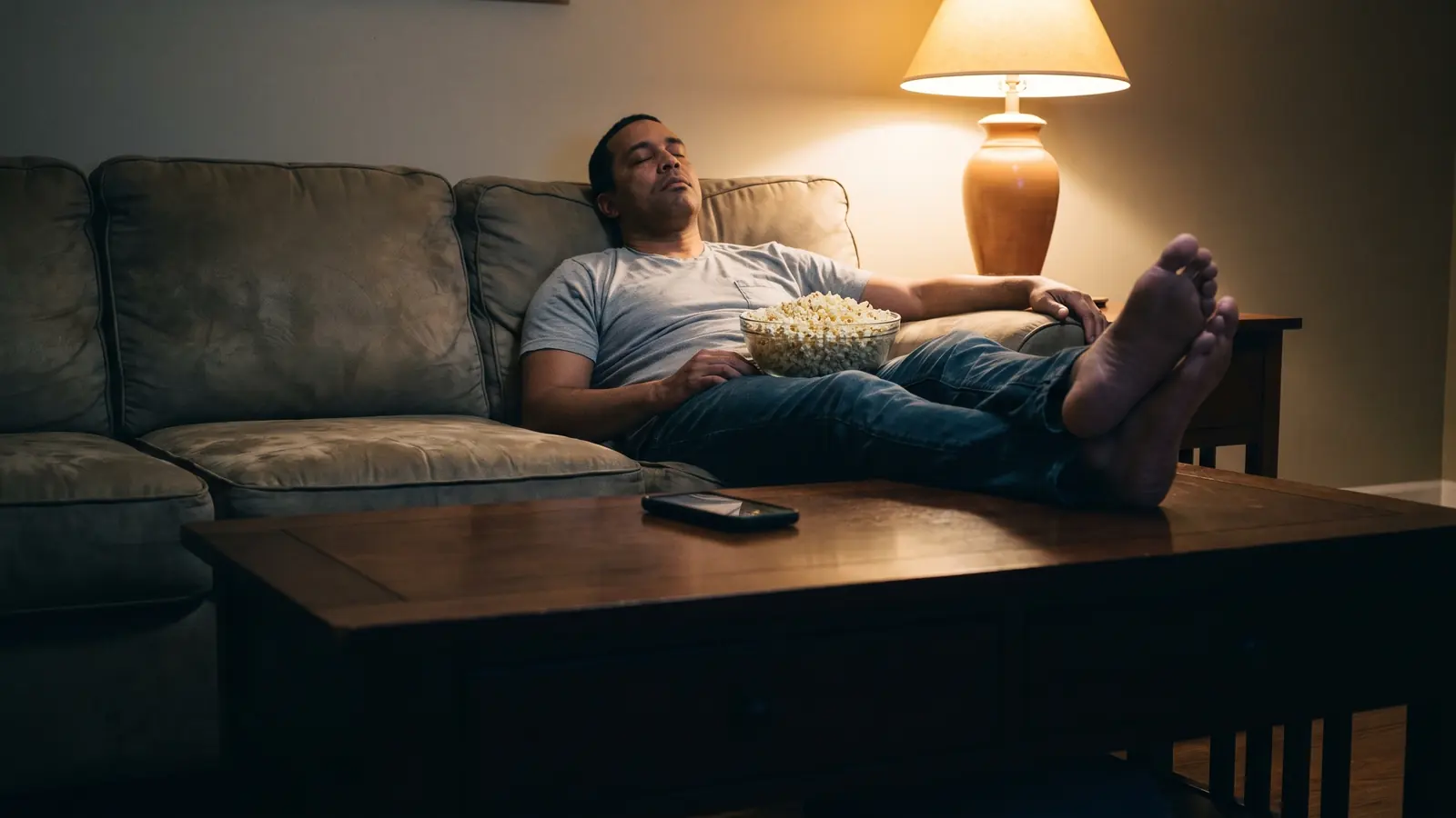 A man asleep on a couch in a dim living room, his phone face-down on the coffee table beside him.
